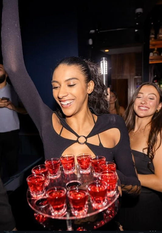 VIP bottle waitress carrying a round shot tray filled with red shots alongside an LED bottle service light baton in a nightclub